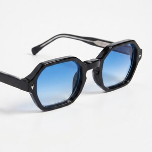 Peut inclure: Lunettes de soleil noires avec une monture géométrique octogonale et des verres dégradés bleus. Les lunettes ont un petit accent argenté sur le devant de la monture. Les branches sont noires avec une rayure blanche.