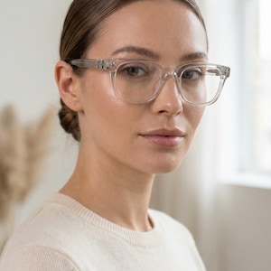 May include: Clear, square-framed eyeglasses with decorative accents on the temples. The glasses are transparent, offering a modern and minimalist aesthetic. The image shows a close-up of the glasses, highlighting their design and clarity.