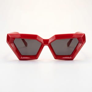 May include: Red geometric sunglasses with dark grey lenses. The sunglasses have a bold, angular frame design. The glasses are set against a white background. These sunglasses are a fashion accessory.