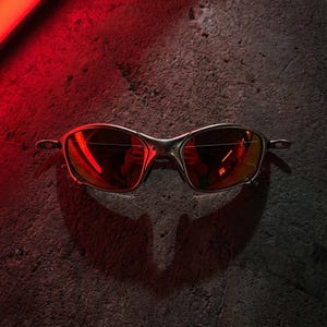 K&ouml;nnte beinhalten: Nahaufnahme einer silberfarbenen Sonnenbrille mit rot get&ouml;nten Gl&auml;sern. Die Brille befindet sich vor einem dunklen, strukturierten Hintergrund, wobei eine rote Lichtquelle einen Schatten wirft. Modernes und elegantes Design.