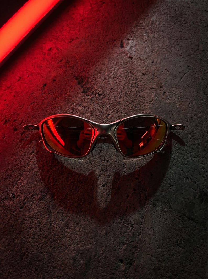 K&ouml;nnte beinhalten: Nahaufnahme einer silberfarbenen Sonnenbrille mit rot get&ouml;nten Gl&auml;sern. Die Brille befindet sich vor einem dunklen, strukturierten Hintergrund, wobei eine rote Lichtquelle einen Schatten wirft. Modernes und elegantes Design.