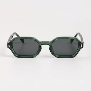 Peut inclure: Lunettes de soleil à monture verte avec verres foncés. Les montures ont une forme géométrique octogonale. Les branches sont d'une nuance de vert plus foncée. Les lunettes sont posées sur un fond blanc.