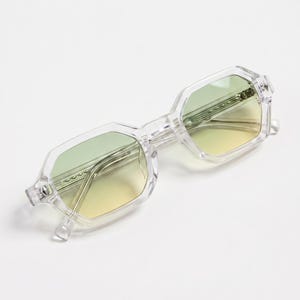 Peut inclure: Lunettes de soleil géométriques transparentes avec des verres dégradés vert-jaune. Les montures sont transparentes et le design est moderne. Les lunettes sont posées sur un fond blanc, mettant en valeur leur forme et leur couleur.