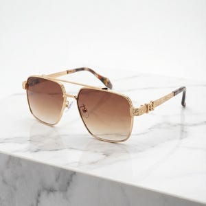 Könnte beinhalten: Goldfarbene Sonnenbrille mit braunen Verlaufsgläsern. Die quadratischen Rahmen haben dekorative Details an den Bügeln und am Steg. Die Bügel haben ein Schildpattmuster in der Nähe der Scharniere. Die Sonnenbrille liegt auf einer weißen Marmoroberfläche.