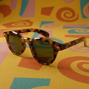 Peut inclure: Lunettes de soleil en écaille de tortue avec des verres foncés, posées sur une surface colorée à motifs. Les lunettes ont une monture arrondie classique et un détail décoratif subtil sur les branches. Le fond présente un design rétro avec des spirales et des formes géométriques.