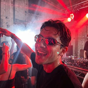 K&ouml;nnte beinhalten: Eine Person mit silberfarbener Sonnenbrille l&auml;chelt breit in einem &uuml;berf&uuml;llten, schwach beleuchteten Raum mit roter Beleuchtung. Die Person tr&auml;gt ein schwarzes T-Shirt und scheint auf einem Konzert oder einer Party zu sein.