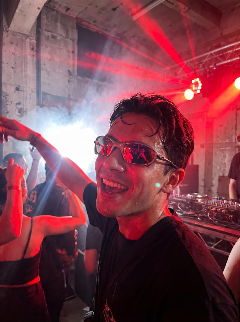 K&ouml;nnte beinhalten: Eine Person mit silberfarbener Sonnenbrille l&auml;chelt breit in einem &uuml;berf&uuml;llten, schwach beleuchteten Raum mit roter Beleuchtung. Die Person tr&auml;gt ein schwarzes T-Shirt und scheint auf einem Konzert oder einer Party zu sein.