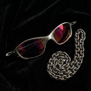 K&ouml;nnte beinhalten: Silberne Sonnenbrille mit rosa get&ouml;nten Gl&auml;sern und eine silberne Halskette auf schwarzem Hintergrund. Die Sonnenbrille hat ein schlankes, modernes Design. Die Kette ist aufwendig detailliert.