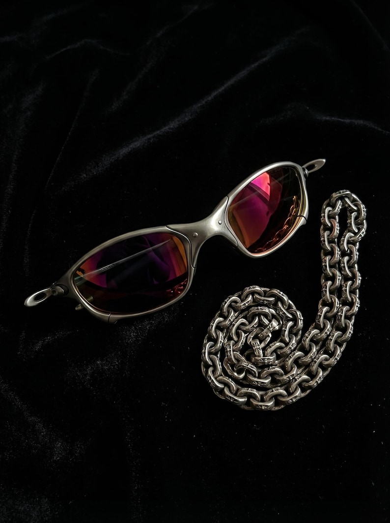 K&ouml;nnte beinhalten: Silberne Sonnenbrille mit rosa get&ouml;nten Gl&auml;sern und eine silberne Halskette auf schwarzem Hintergrund. Die Sonnenbrille hat ein schlankes, modernes Design. Die Kette ist aufwendig detailliert.