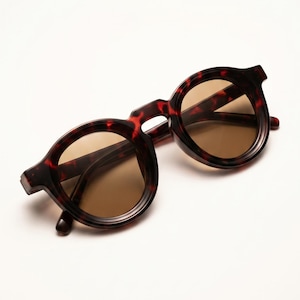 May include: Round-framed sunglasses with a tortoiseshell pattern. The frames are a mix of dark brown and reddish-brown hues, with a subtle, textured appearance. The lenses are a warm, light brown color, providing a contrast to the darker frames. The sunglasses are set against a white background.