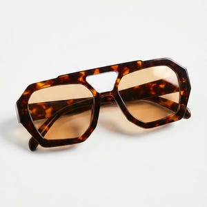 May include: Geometric tortoiseshell sunglasses. The frames are dark brown with amber lenses. The sunglasses have a distinctive shape with a bridge cutout. The lenses are a warm amber colour.