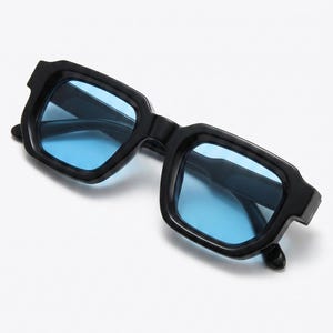 Può includere: Occhiali da sole quadrati neri con lenti azzurre. Gli occhiali da sole hanno un design con montatura spessa e audace. Le lenti sono di un colore blu brillante e uniforme. Gli occhiali sono su uno sfondo bianco.