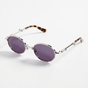 Peut inclure: Lunettes de soleil ovales avec une monture en métal argenté et des verres violets foncés. Les branches présentent un motif décoratif et des extrémités en écaille de tortue. Un accessoire élégant.