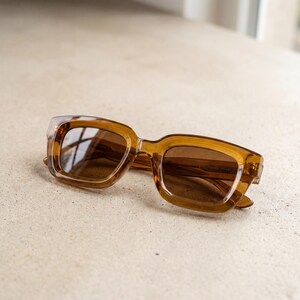 Peut inclure: Lunettes de soleil carrées marron avec verres teintés. Les lunettes ont une monture épaisse et translucide et reposent sur une surface de couleur claire. Les verres reflètent l'environnement.