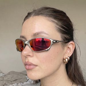 K&ouml;nnte beinhalten: Silberfarbene Sonnenbrille mit roten Gl&auml;sern. Die sportliche Brille hat ein umlaufendes Design und wird von einer Person getragen. Die Gl&auml;ser reflektieren die Umgebung. Goldene Creolen und eine goldene Kette sind ebenfalls zu sehen.