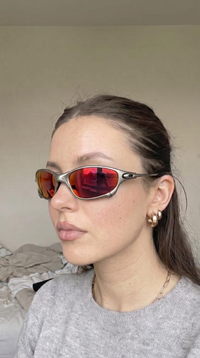K&ouml;nnte beinhalten: Silberfarbene Sonnenbrille mit roten Gl&auml;sern. Die sportliche Brille hat ein umlaufendes Design und wird von einer Person getragen. Die Gl&auml;ser reflektieren die Umgebung. Goldene Creolen und eine goldene Kette sind ebenfalls zu sehen.