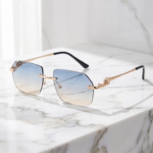 Peut inclure: Lunettes de soleil sans monture avec verres dégradés bleus et beiges. Les montures sont dorées avec des branches noires. Les lunettes de soleil reposent sur une surface en marbre blanc. Le style général est moderne et à la mode.