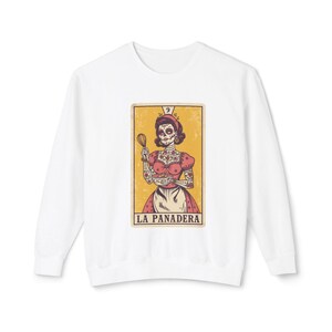 Könnte beinhalten: Weißes Sweatshirt mit Tarotkarten-Design. Die Karte zeigt eine Skelettfrau in einem rosa Kleid mit Schneebesen und dem Text "LA PANADERA" unten. Die Karte hat einen gelben Hintergrund und einen Vintage-Look.