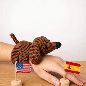 Peut inclure: Un jouet teckel en crochet marron en forme de manchette, doté d'un nez et d'yeux noirs. Deux petits drapeaux en tissu sont montés sur des socles en bois ronds : l'un représentant les États-Unis et l'autre le drapeau national espagnol.