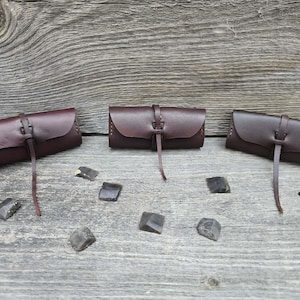 May include: Three handcrafted leather pouches in varying shades of brown, each with a flap closure and a leather strap. The pouches are arranged on a weathered wooden surface, with several dark, irregularly shaped stones scattered around them.