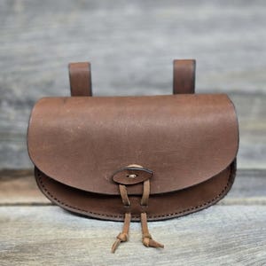 May include: A brown leather pouch with a rounded base and a flap closure. The pouch features two belt loops at the back and a leather cord with tassels. The leather has a slightly worn appearance.