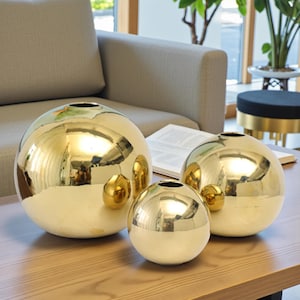 Op de afbeelding: Drie goudkleurige, bolvormige vazen van verschillende grootte staan op een houten tafel. De vazen hebben een gepolijst, reflecterend oppervlak met een kleine opening aan de bovenkant. Naast elke vaas staat een kleine gouden bol.
