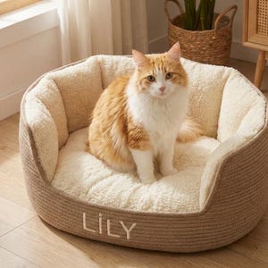 以下が含まれることがあります： ふわふわのインナーと織りのアウターを備えた、ベージュとクリーム色の猫用ベッド。前面には「LILY」という名前が刺繍されています。オレンジと白の猫がベッドの中に座っています。