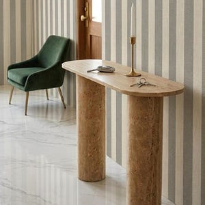 Puede incluir: Una consola de mármol beige con dos patas cilíndricas. Un candelabro dorado con una vela blanca está sobre la mesa, junto con llaves y un llavero de coche. Una silla de terciopelo verde y papel pintado a rayas están en el fondo.
