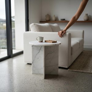 Puede incluir: Una mesa auxiliar de m&aacute;rmol blanco con una tapa redonda y un dise&ntilde;o de base &uacute;nico. Una taza peque&ntilde;a y una bandeja de madera est&aacute;n sobre la mesa. La mesa est&aacute; junto a un sof&aacute; blanco en un sal&oacute;n moderno.