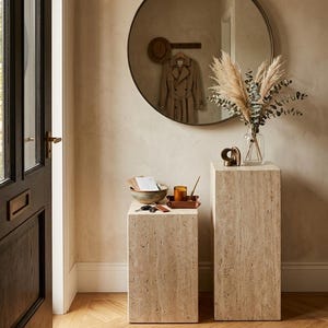 May include: Two rectangular travertine pedestals with a porous, beige, natural stone texture. The shorter pedestal holds a ceramic bowl, a small tray with a candle, and keys. The taller pedestal supports a clear glass vase with dried pampas grass.