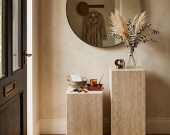 Puede incluir: Dos pedestales rectangulares de travertino con una textura de piedra natural beige y porosa. El pedestal más bajo sostiene un cuenco de cerámica, una bandeja pequeña con una vela y llaves. El más alto sostiene un jarrón de cristal con hierbas.