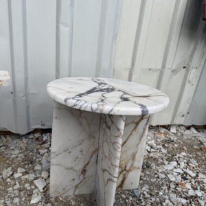 Puede incluir: Una mesa auxiliar de m&aacute;rmol blanco con una tapa redonda y un dise&ntilde;o de base &uacute;nico. La mesa presenta vetas grises oscuras y marrones en todo el m&aacute;rmol. Una pieza de acento elegante para cualquier habitaci&oacute;n.