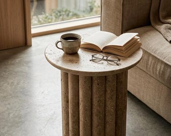 Puede incluir: Una pequeña mesa auxiliar redonda con una parte superior beige claro texturizada y una base hecha de columnas cilíndricas verticales. Una taza de café, un libro abierto y gafas descansan sobre la mesa. Un sofá beige y una ventana están en el fondo.