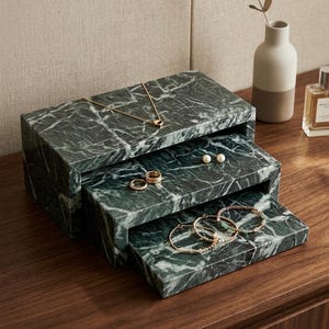 May include: A dark green marble jewelry organizer featuring two stacked, rectangular tiers. The top tier holds a gold chain necklace, while the lower sliding tier displays two gold rings, a pair of pearl stud earrings, and three beaded bracelets.