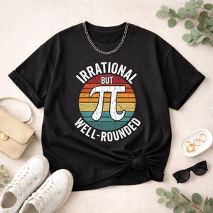 Peut inclure: T-shirt noir avec le texte "IRRATIONAL BUT WELL-ROUNDED" dans un design rétro avec le symbole Pi. Le motif est placé sur un fond aux couleurs du coucher de soleil. Un collier en chaîne argentée est drapé sur le t-shirt.