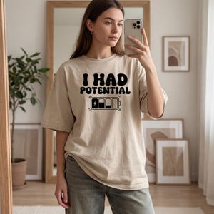 Può includere: T-shirt beige con la scritta nera "I HAD POTENTIAL" e un'icona della batteria. La maglietta ha una vestibilità ampia e maniche corte. La grafica è centrata sul davanti.