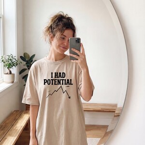 以下が含まれることがあります： 「I HAD POTENTIAL」の黒い文字と下降グラフがプリントされたベージュ色のTシャツ。カジュアルでオーバーサイズのスタイルです。鏡の前で撮影された画像。