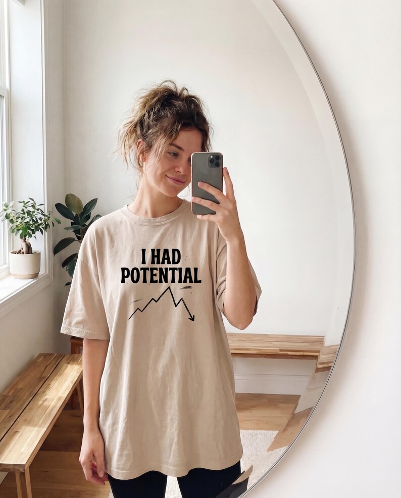 以下が含まれることがあります： 「I HAD POTENTIAL」の黒い文字と下降グラフがプリントされたベージュ色のTシャツ。カジュアルでオーバーサイズのスタイルです。鏡の前で撮影された画像。