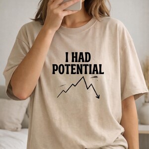 以下が含まれることがあります： 「I HAD POTENTIAL」の黒い文字と下降グラフがプリントされたベージュ色のTシャツ。ニュートラルカラーで半袖です。