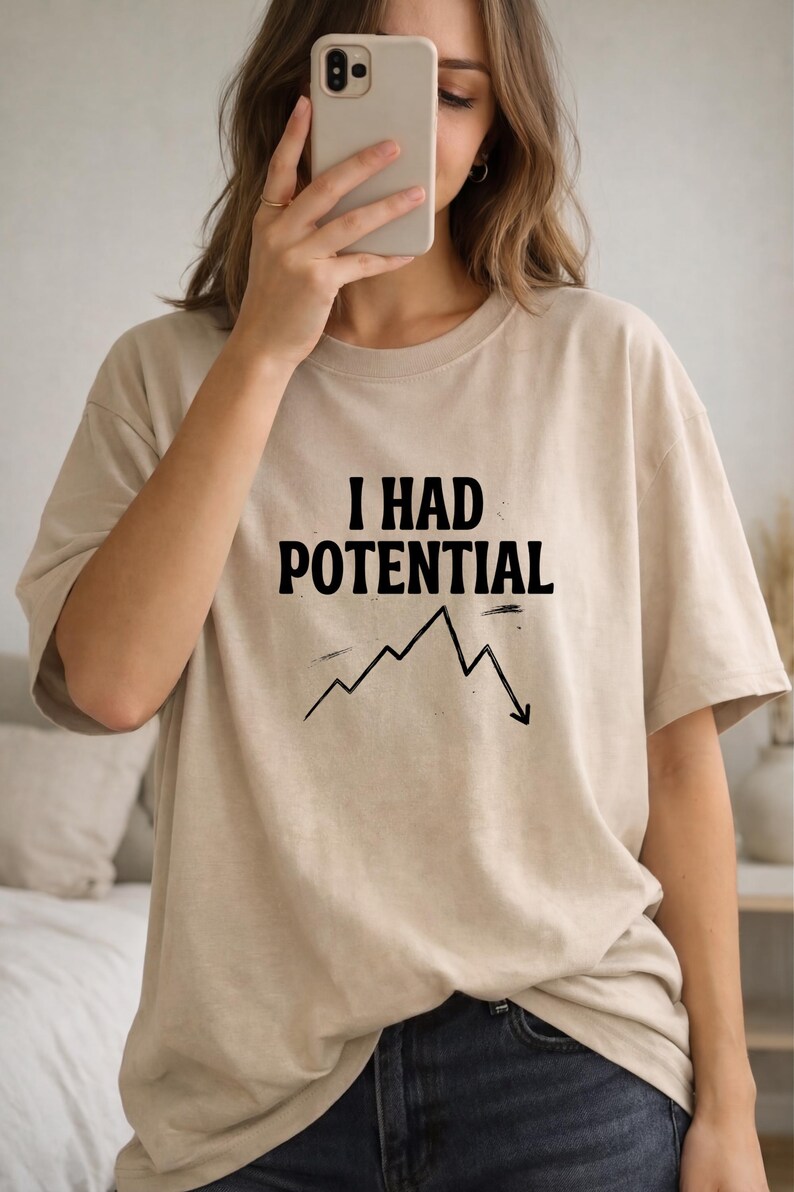 以下が含まれることがあります： 「I HAD POTENTIAL」の黒い文字と下降グラフがプリントされたベージュ色のTシャツ。ニュートラルカラーで半袖です。