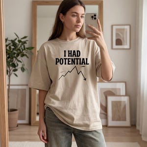 以下が含まれることがあります： 「I HAD POTENTIAL」の黒い文字と下降グラフがプリントされたベージュのオーバーサイズTシャツ。グレーのジーンズと合わせています。鏡の前で撮影された画像です。