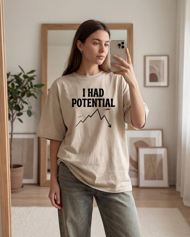 以下が含まれることがあります： 「I HAD POTENTIAL」の黒い文字と下降グラフがプリントされたベージュのオーバーサイズTシャツ。グレーのジーンズと合わせています。鏡の前で撮影された画像です。