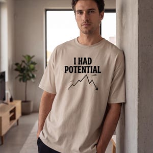 以下が含まれることがあります： ベージュ色の半袖Tシャツ。黒の太字で「I HAD POTENTIAL」の文字がプリントされています。その下に、下降傾向を示す黒い折れ線グラフがあります。ゆったりとしたフィット感のTシャツです。