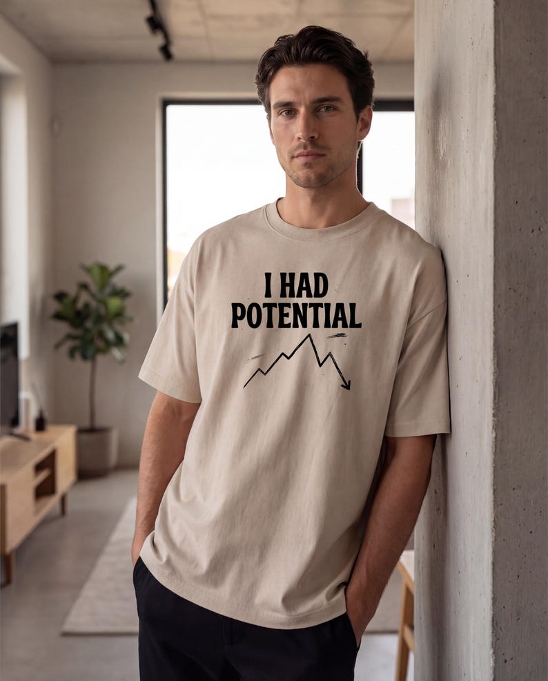 以下が含まれることがあります： ベージュ色の半袖Tシャツ。黒の太字で「I HAD POTENTIAL」の文字がプリントされています。その下に、下降傾向を示す黒い折れ線グラフがあります。ゆったりとしたフィット感のTシャツです。