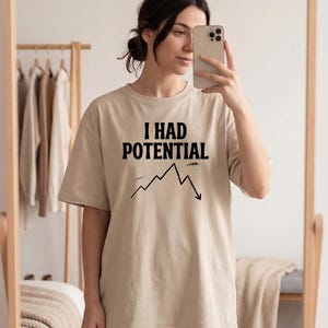 以下が含まれることがあります： 「I HAD POTENTIAL」の黒い文字と、下降傾向の折れ線グラフがプリントされたベージュ色のTシャツ。半袖でカジュアルなスタイルです。