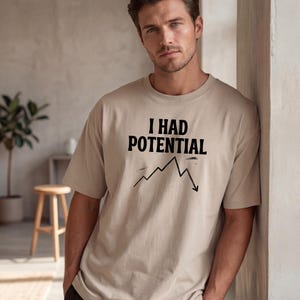 以下が含まれることがあります： 「I HAD POTENTIAL」の黒い文字と下降グラフがプリントされたベージュ色のTシャツ。ニュートラルカラーでリラックスしたフィット感です。