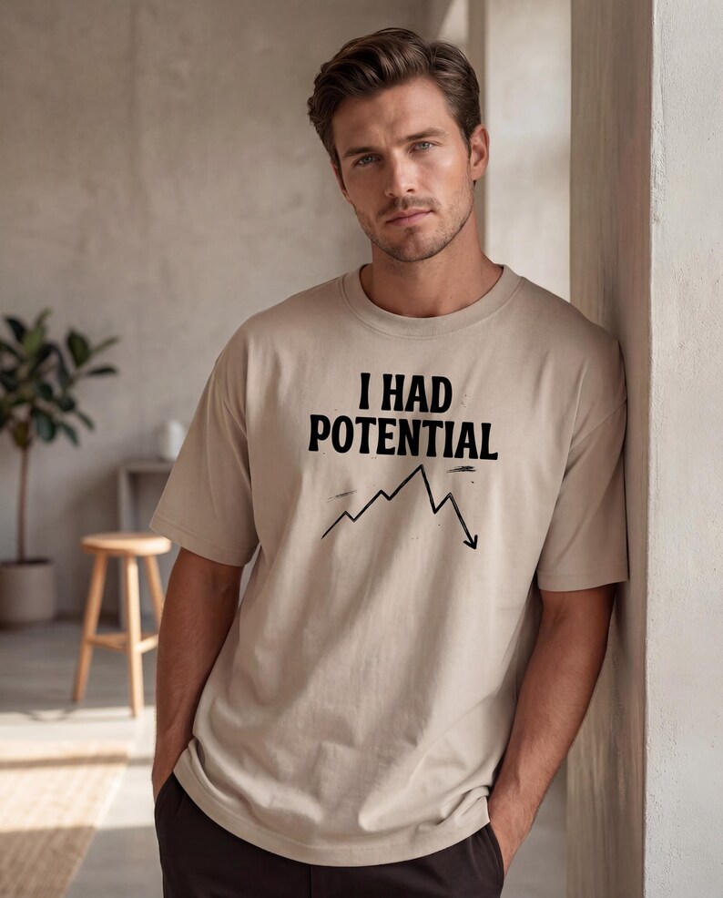 以下が含まれることがあります： 「I HAD POTENTIAL」の黒い文字と下降グラフがプリントされたベージュ色のTシャツ。ニュートラルカラーでリラックスしたフィット感です。