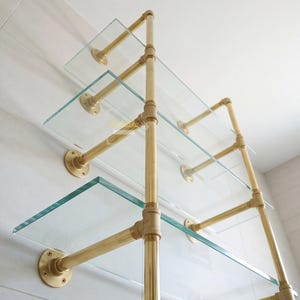 May include: A wall-mounted shelving unit featuring three clear glass shelves supported by a brass-toned metal pipe frame. The structure includes vertical pipes, horizontal pipe arms, and circular wall flanges, all finished in a polished gold tone.