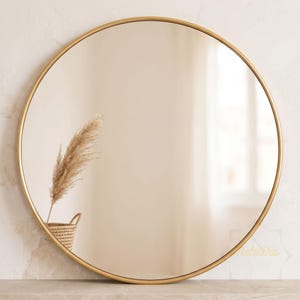 May include: A large circular wall mirror featuring a thin, polished gold-toned metal frame. The reflective glass surface shows a clear view of a woven basket containing a tall, feathery pampas grass stem, creating a soft and neutral reflection.