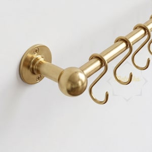 May include: A brushed gold metal wall-mounted rail featuring a cylindrical rod with a spherical end cap and a circular mounting plate secured by two screws. Several matching S-shaped hooks are suspended from the horizontal bar for hanging items.